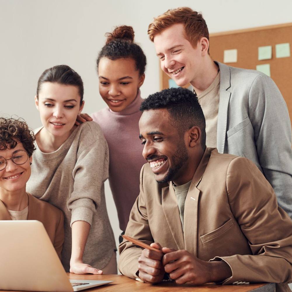 A smiling team of colleagues gathered around a laptop in a bright office